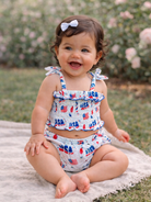 Smiling baby in patriotic outfit sitting on a blanket with roses in the background.