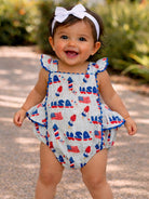 Smiling baby in a patterned outfit with US flags, enjoying the outdoors.