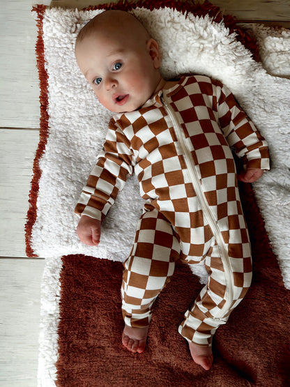 Infant lying on a soft rug, wearing a brown and white checkered zip-up onesie, with a curious expression.