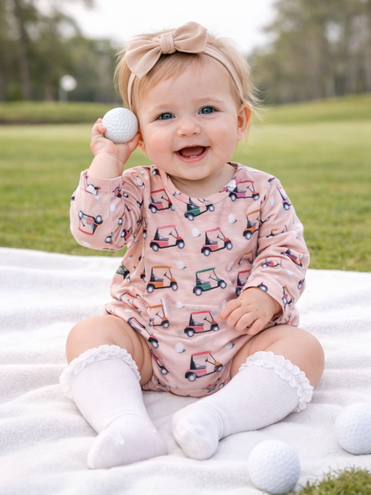 Smiling baby sitting on a blanket, holding a golf ball, wearing a pink outfit and white stockings outdoors.