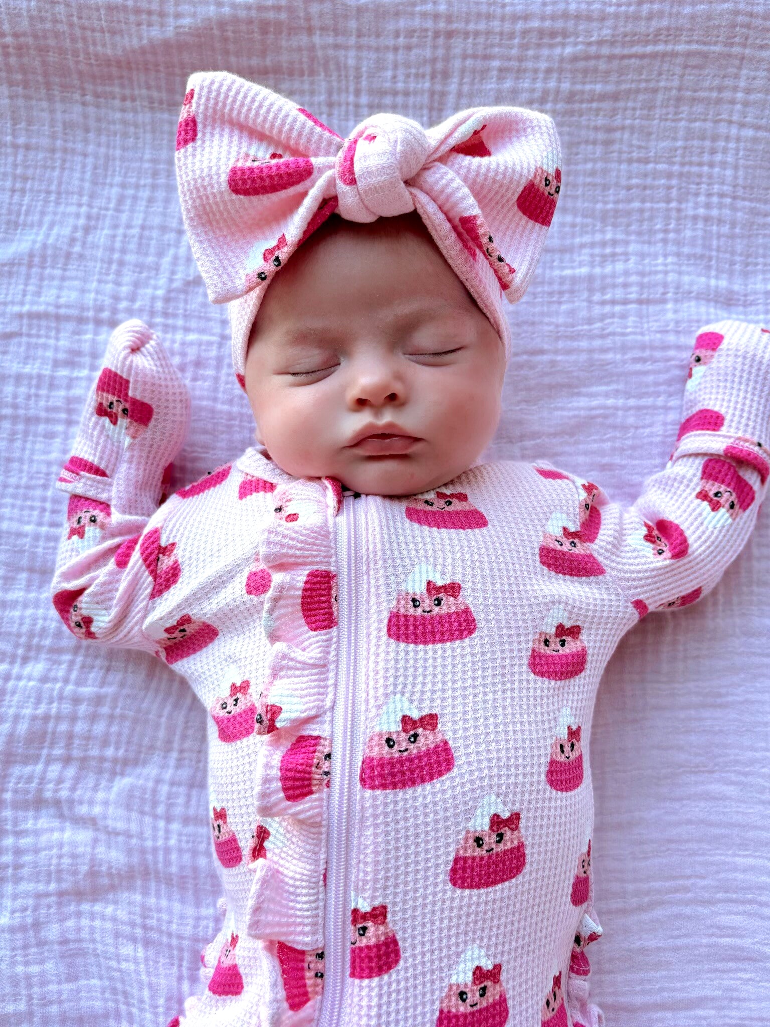 Sleeping baby in a pink outfit with a bow, surrounded by soft textures and pastel cupcake patterns.