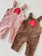 Two pairs of toddler corduroy overalls, one pink with a barn patch, and one brown, laid flat on a light surface.