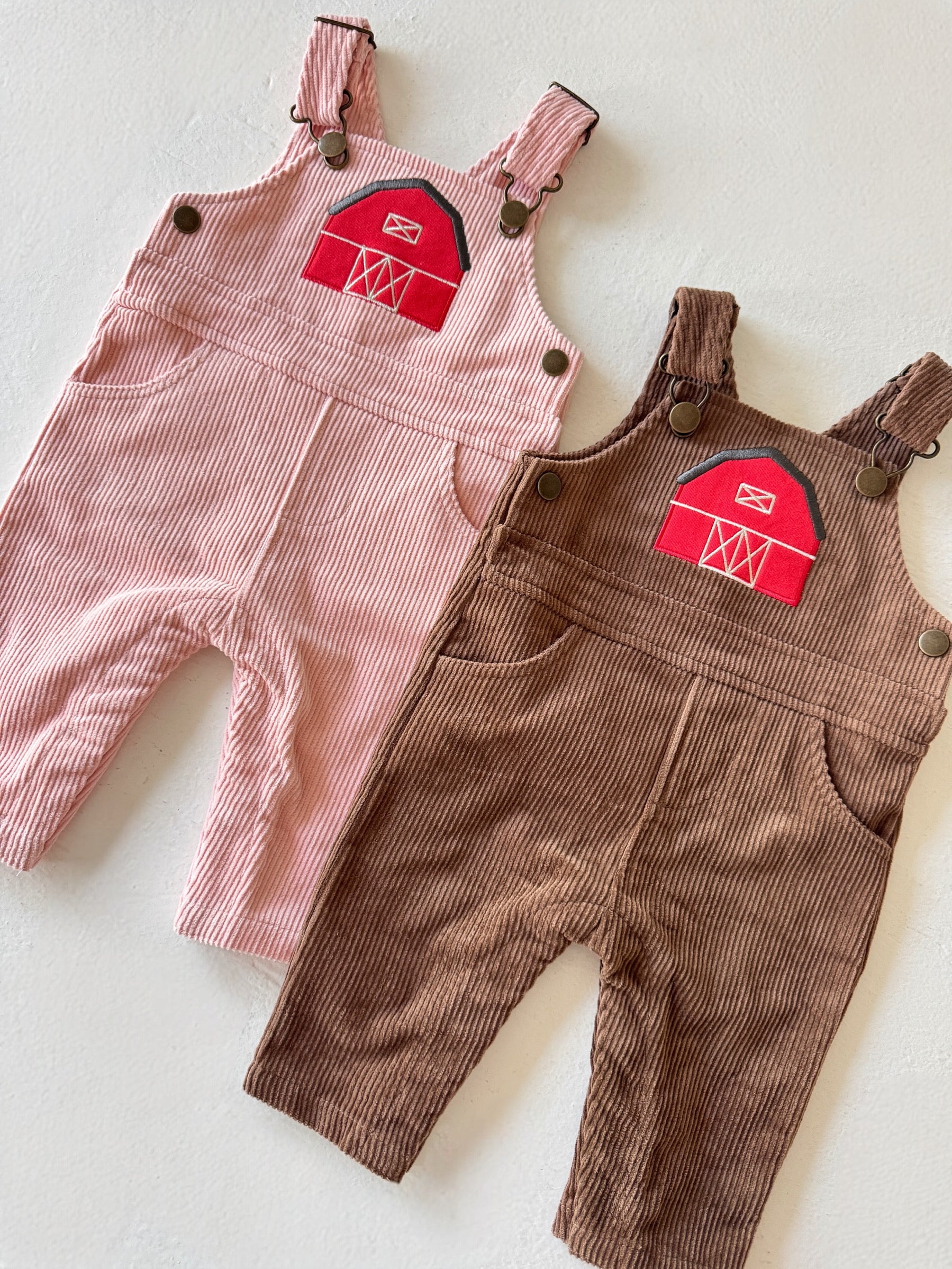 Two pairs of toddler corduroy overalls, one pink with a barn patch, and one brown, laid flat on a light surface.