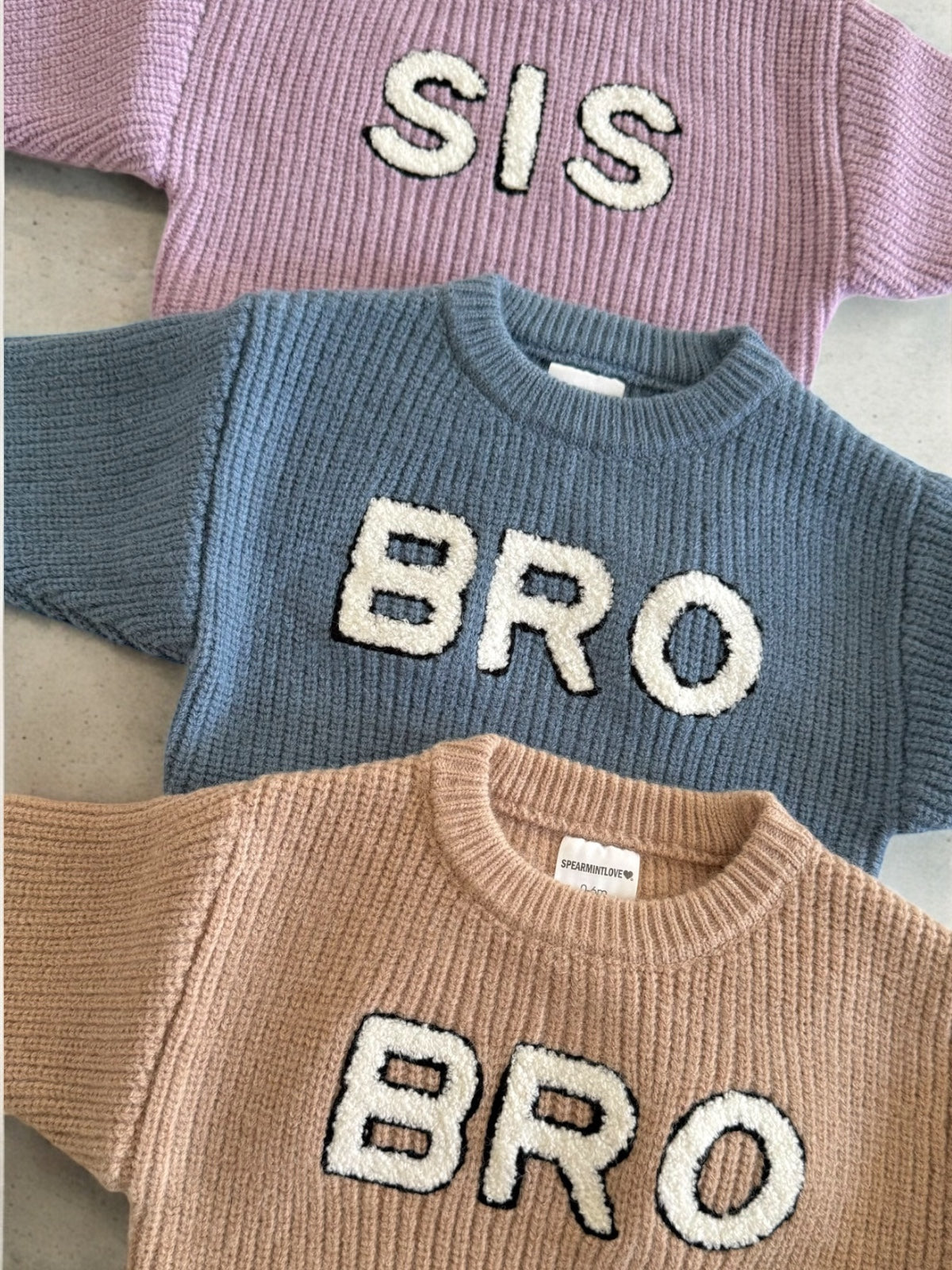 Three knitted sweaters in pastel colors, featuring the words "SIS" and "BRO" in bold, textured letters.