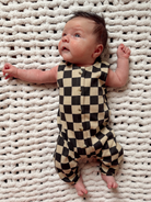 A baby in a black and beige checkered outfit lies on a textured white blanket, looking up.