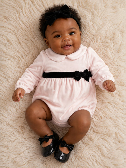 Smiling baby in a pink romper with a black bow on a soft, textured surface.