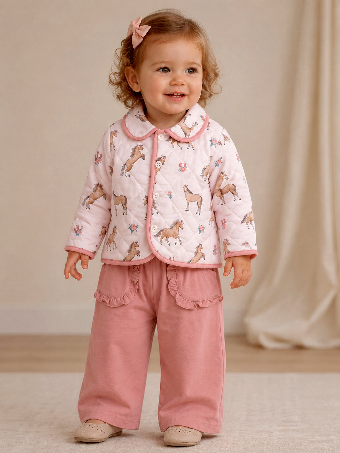 Toddler in a pink horse-patterned jacket and pink pants, smiling with a bow in her hair.