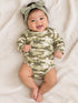 Smiling baby wearing a camouflage onesie and bow headband, lying on a soft, light-colored blanket.