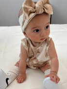 Baby in a beige outfit with a large bow, sitting on a white surface, smiling and reaching for a toy.