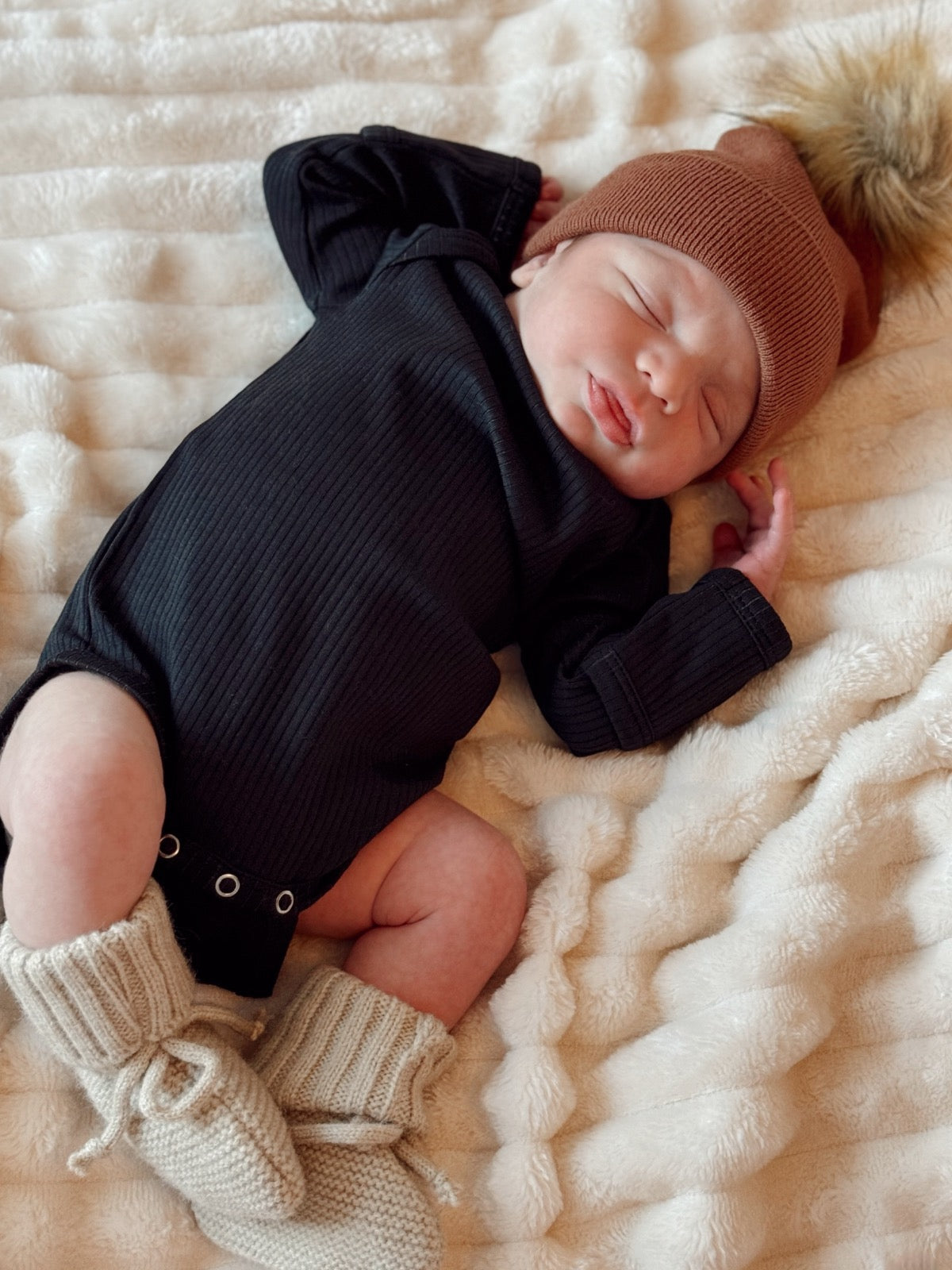 Newborn baby in a black onesie and brown beanie, sleeping on a soft, textured blanket.