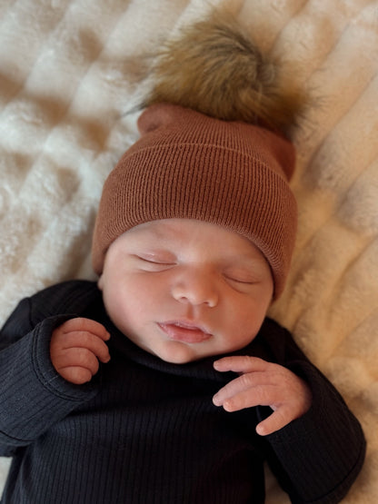Newborn baby sleeping peacefully in a brown beanie with a fluffy pom-pom, wearing a black long-sleeve outfit.
