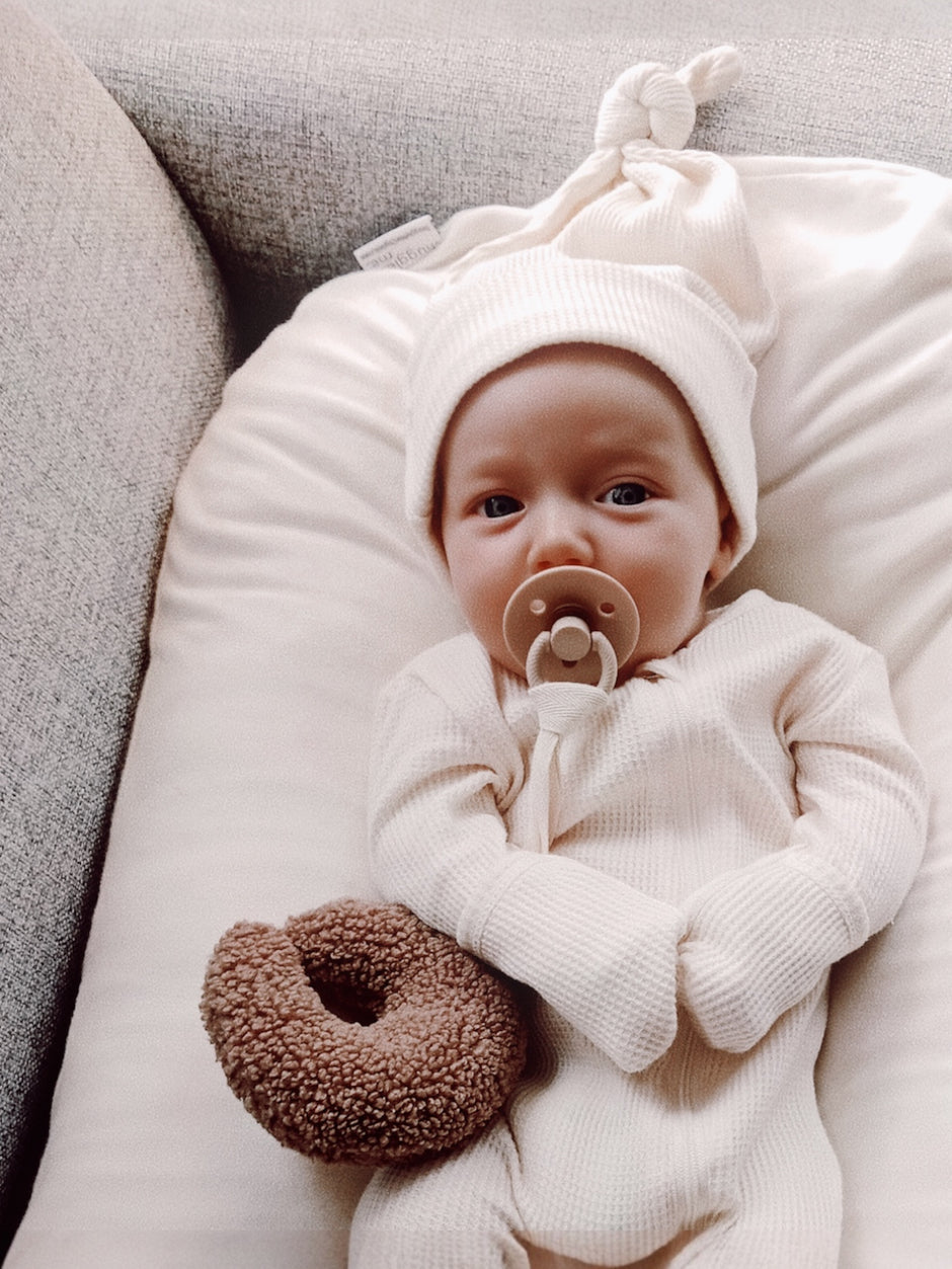 Baby in a cream outfit and hat, holding a soft donut toy and pacifier on a cozy pillow.