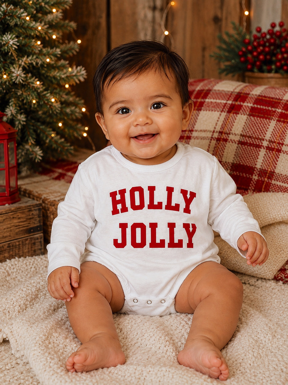 Smiling baby in white "Holly Jolly" onesie, sitting on a cozy blanket with holiday decor in the background.