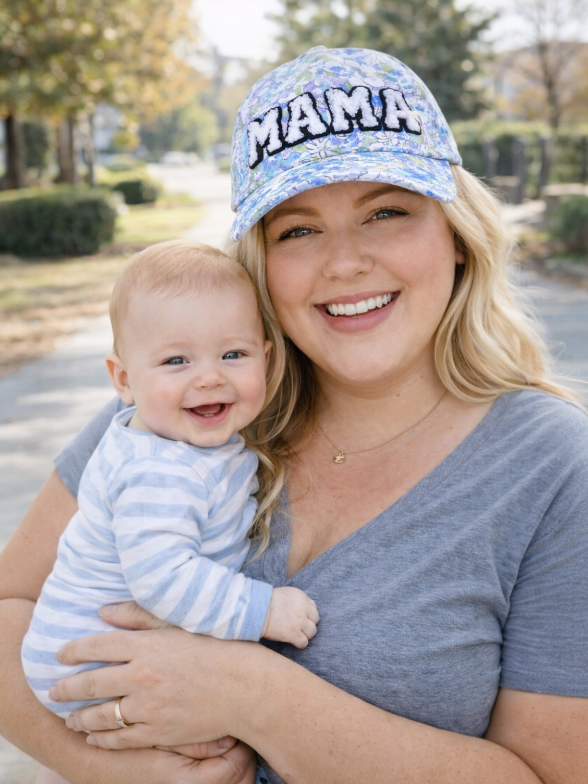 Smiling mother holding a baby outdoors, wearing a blue floral cap with "MAMA" written on it.