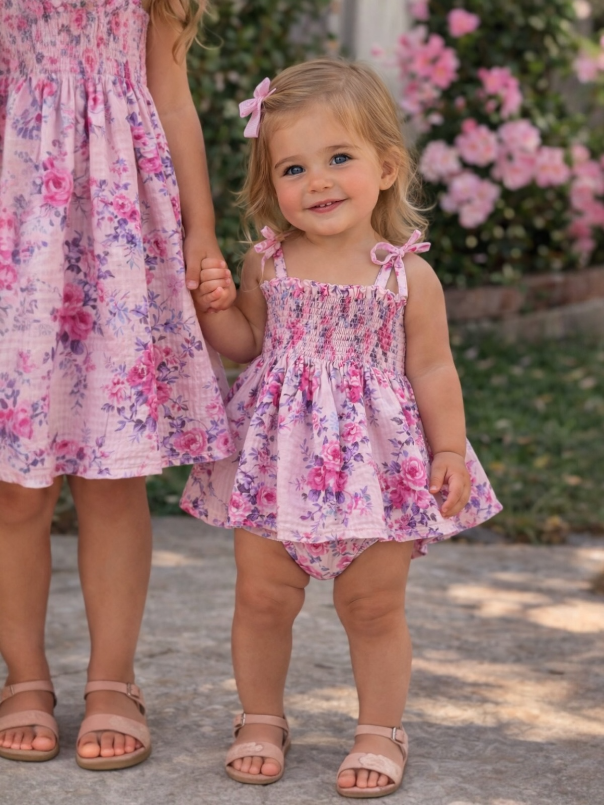 Two young girls hold hands, smiling, wearing matching pink floral dresses in a garden setting.