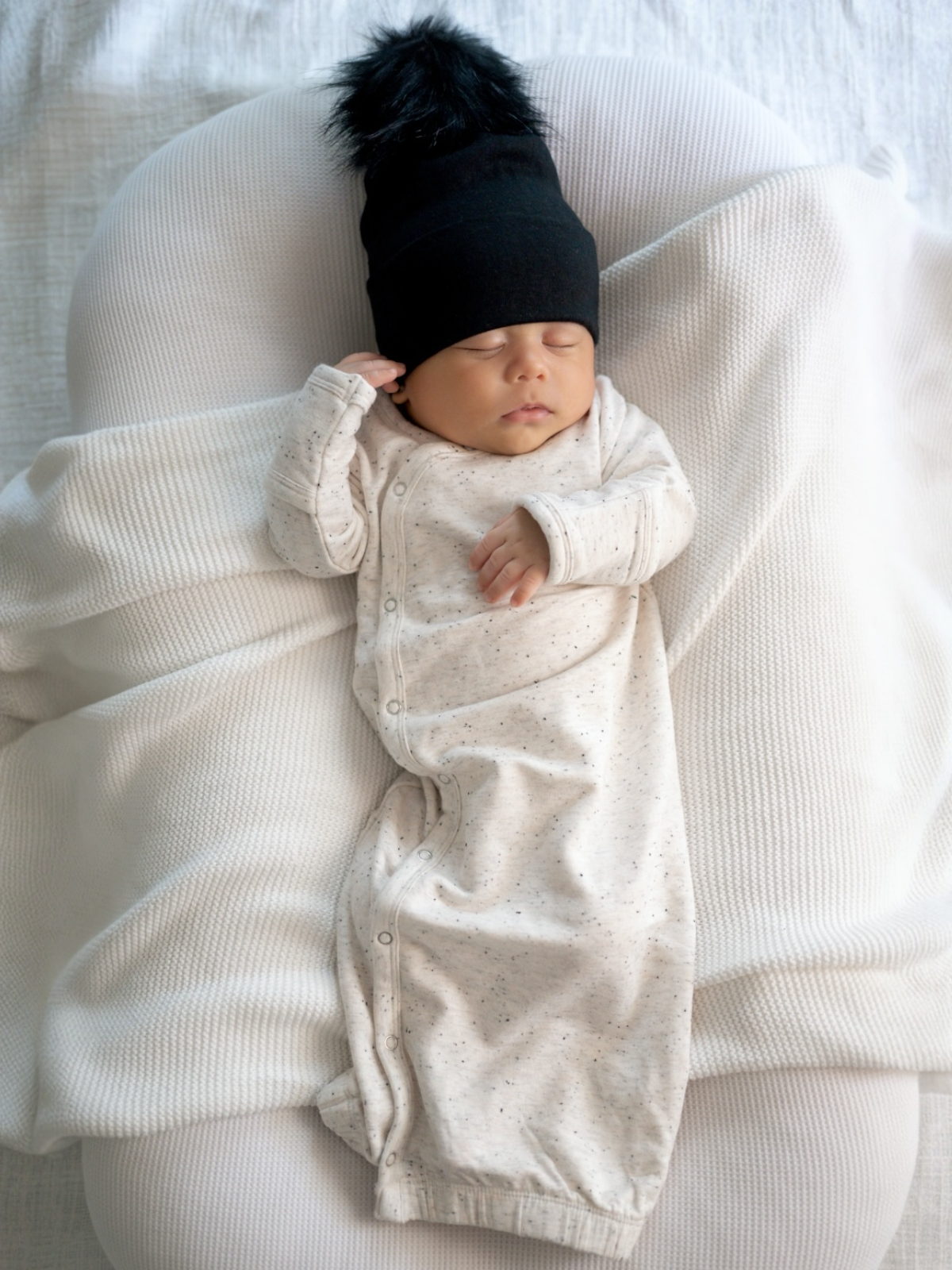 Sleeping baby in a black beanie, wearing a light speckled onesie, on a cozy white blanket.