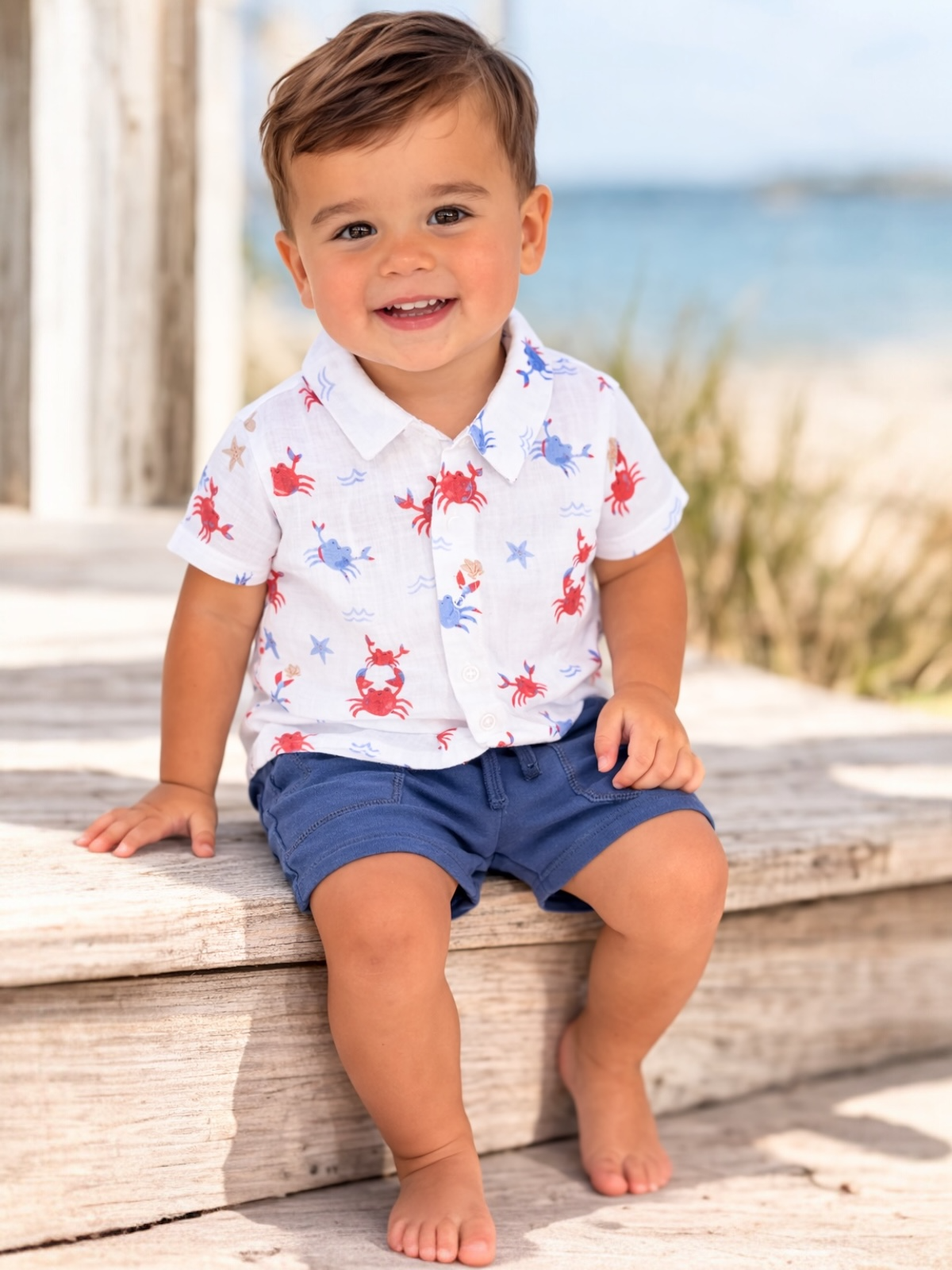 Smiling toddler on a wooden bench, wearing a crab-patterned shirt and blue shorts, with a beach in the background.
