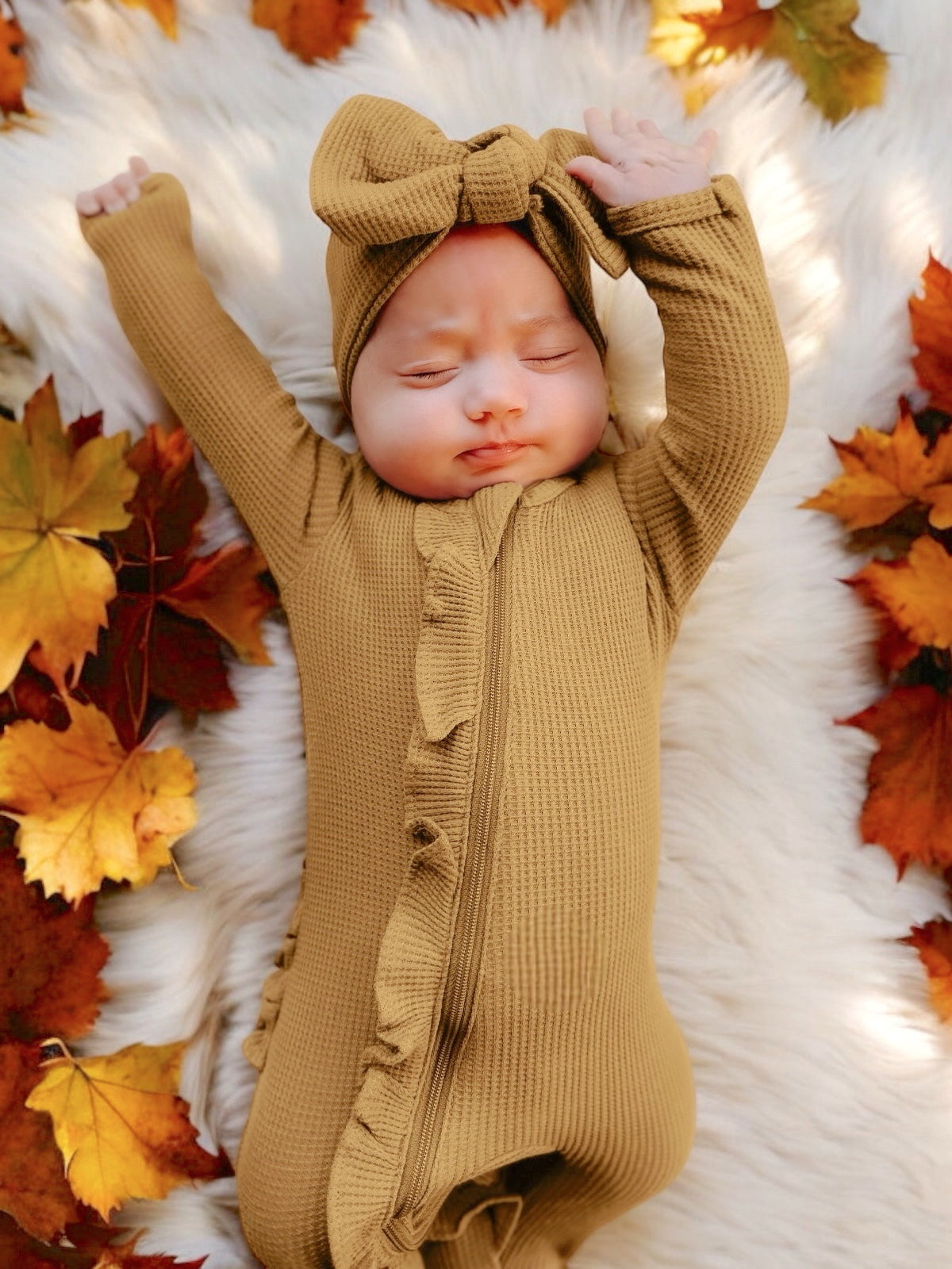 Baby in a mustard outfit and bow lays on a white fur rug surrounded by colorful autumn leaves.