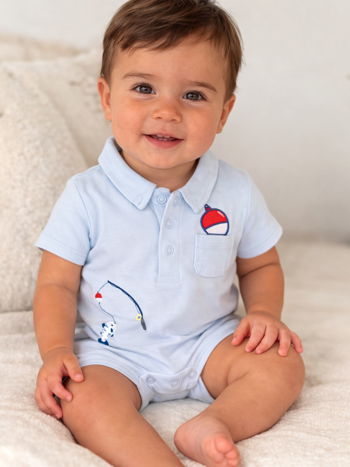 Smiling toddler in a light blue polo onesie with fishing-themed embroidery, seated on a fluffy white surface.
