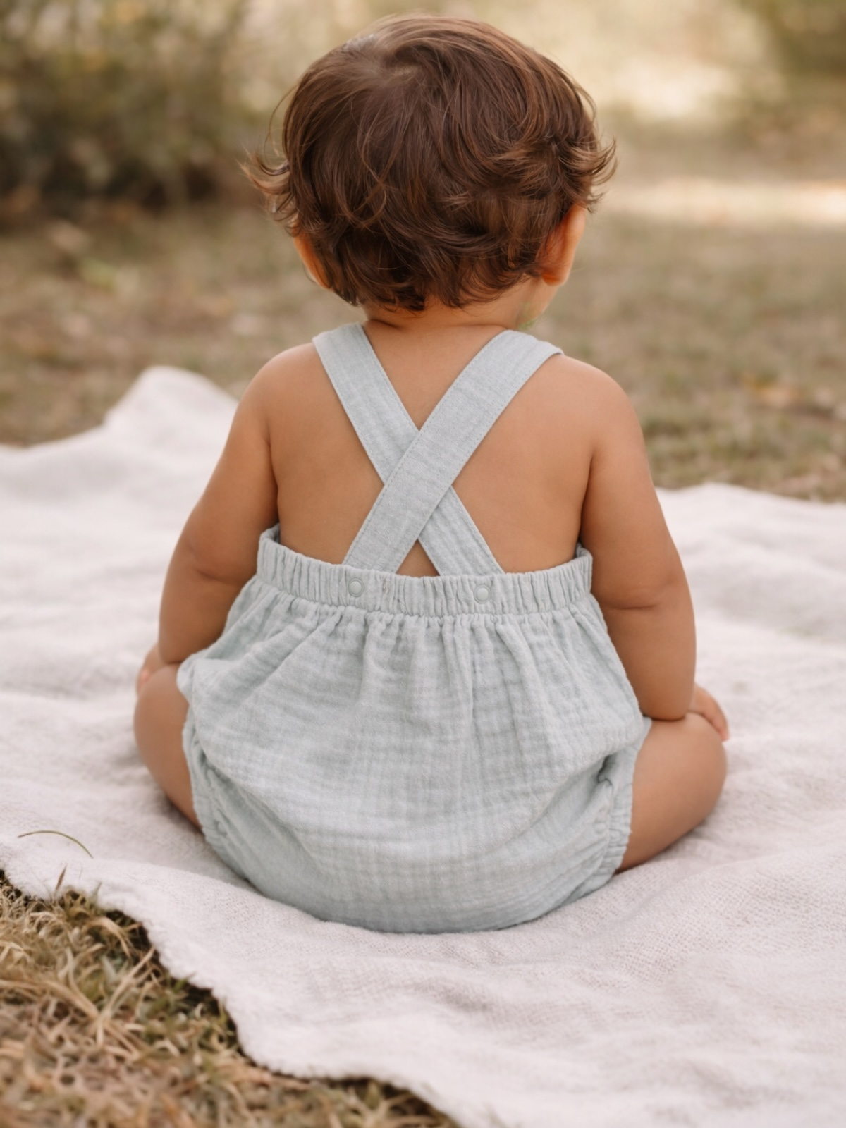 Toddler in a light blue outfit sits on a blanket, viewed from behind, with soft grass and blurred greenery in the background.