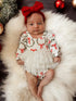 Baby girl in festive outfit with red bows, lying on soft white fur surrounded by holiday decorations.