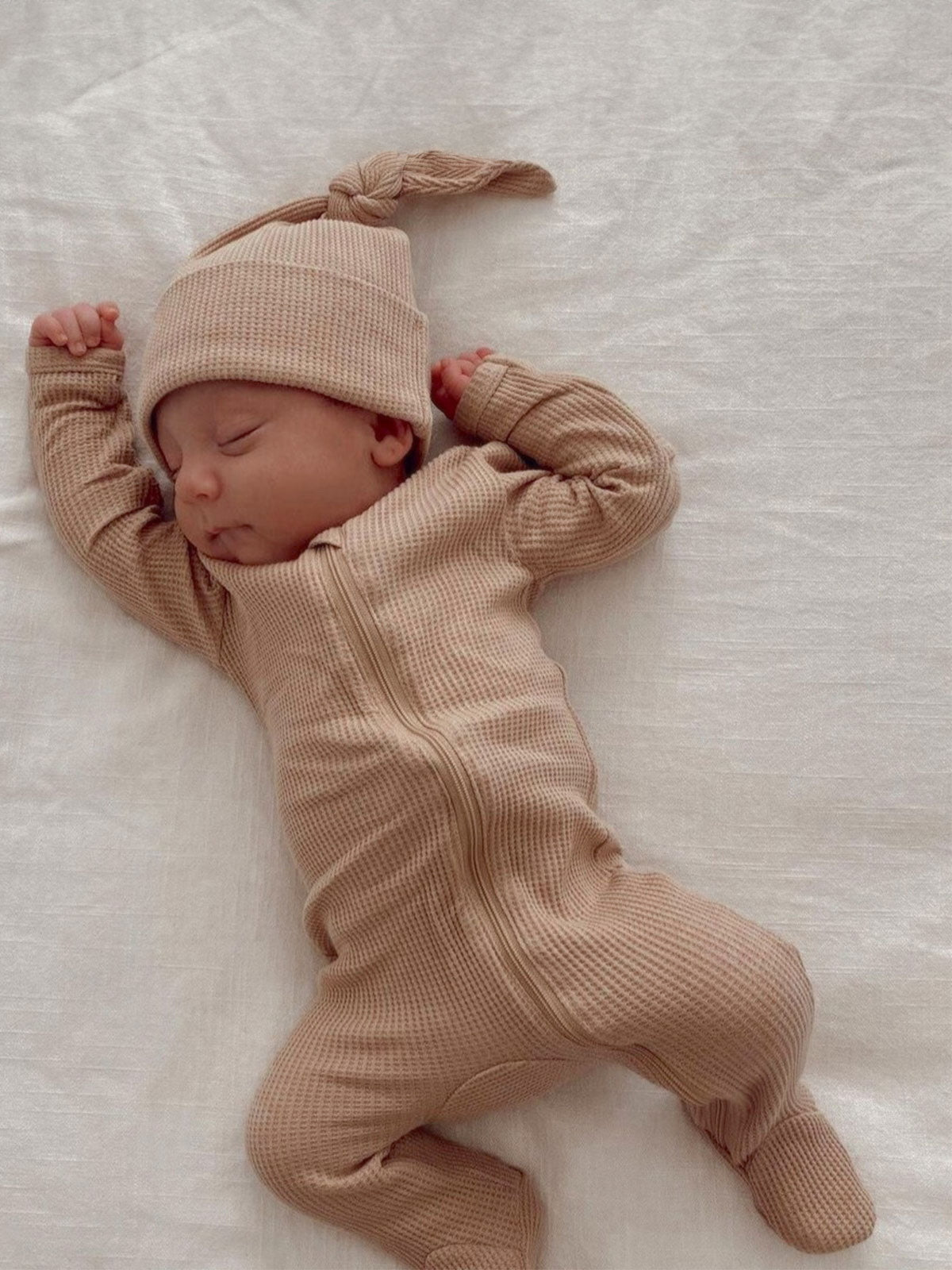 Sleeping newborn dressed in a beige knit onesie and hat, lying on a soft white blanket.