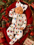 Baby in festive outfit with Christmas lights, sleeping peacefully on a red blanket surrounded by greenery and holiday decor.