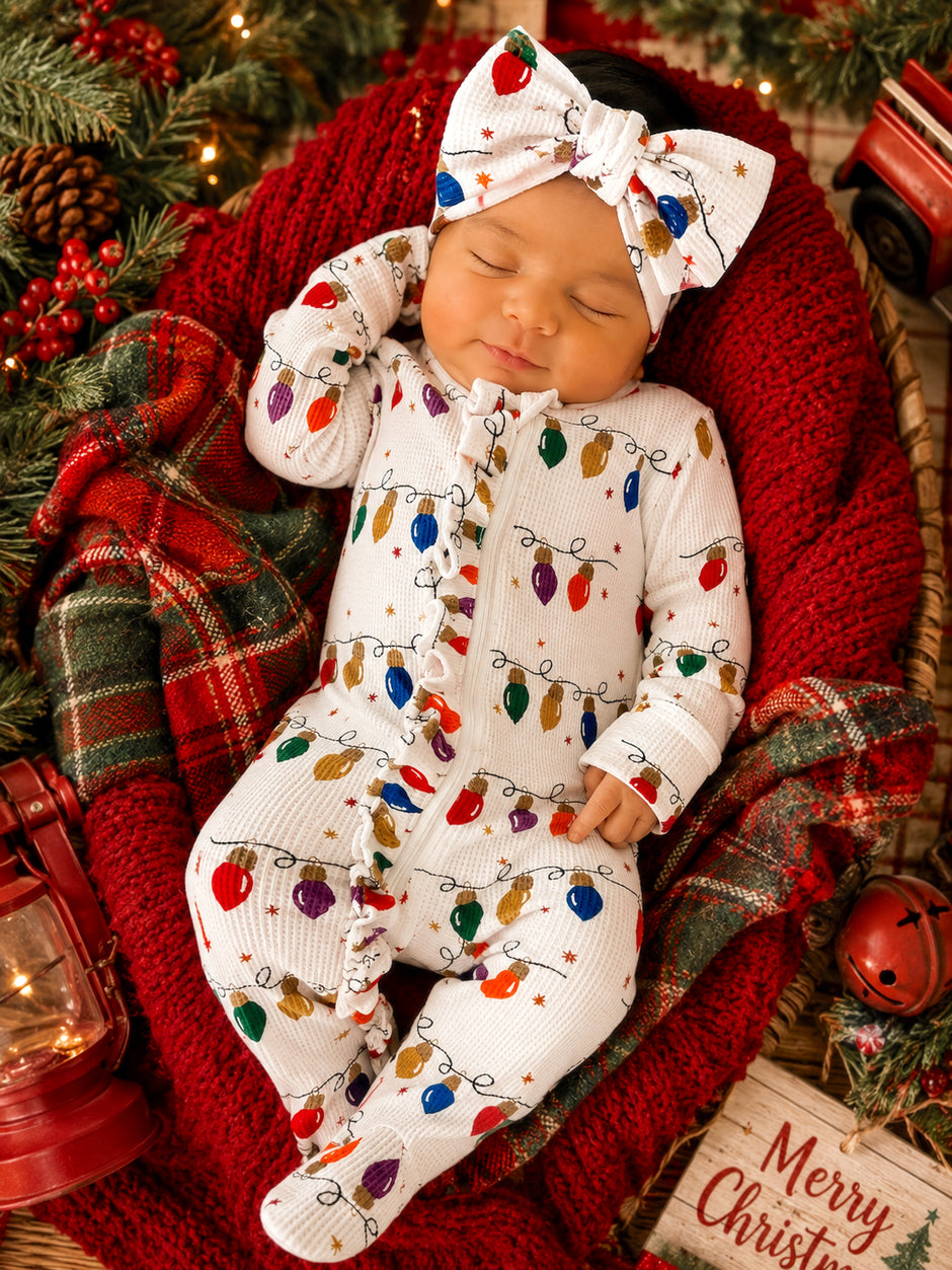 Baby in festive outfit with Christmas lights, sleeping peacefully on a red blanket surrounded by greenery and holiday decor.