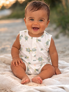 Smiling baby in a turtle-patterned onesie sitting on a sandy beach, enjoying a sunny day.