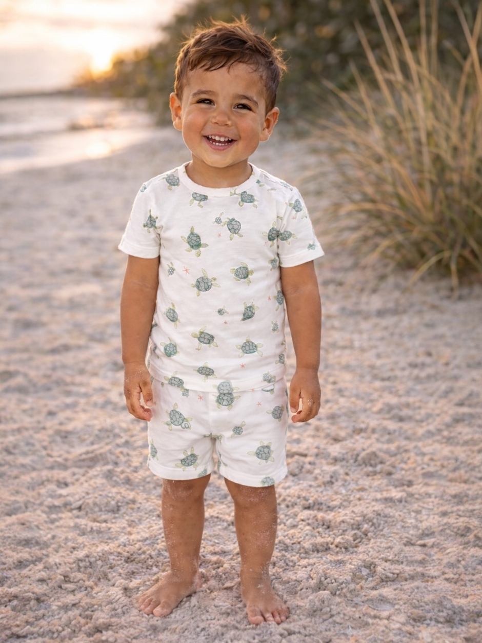 Smiling child in turtle-patterned outfit standing barefoot on sandy beach with soft sunlight in the background.