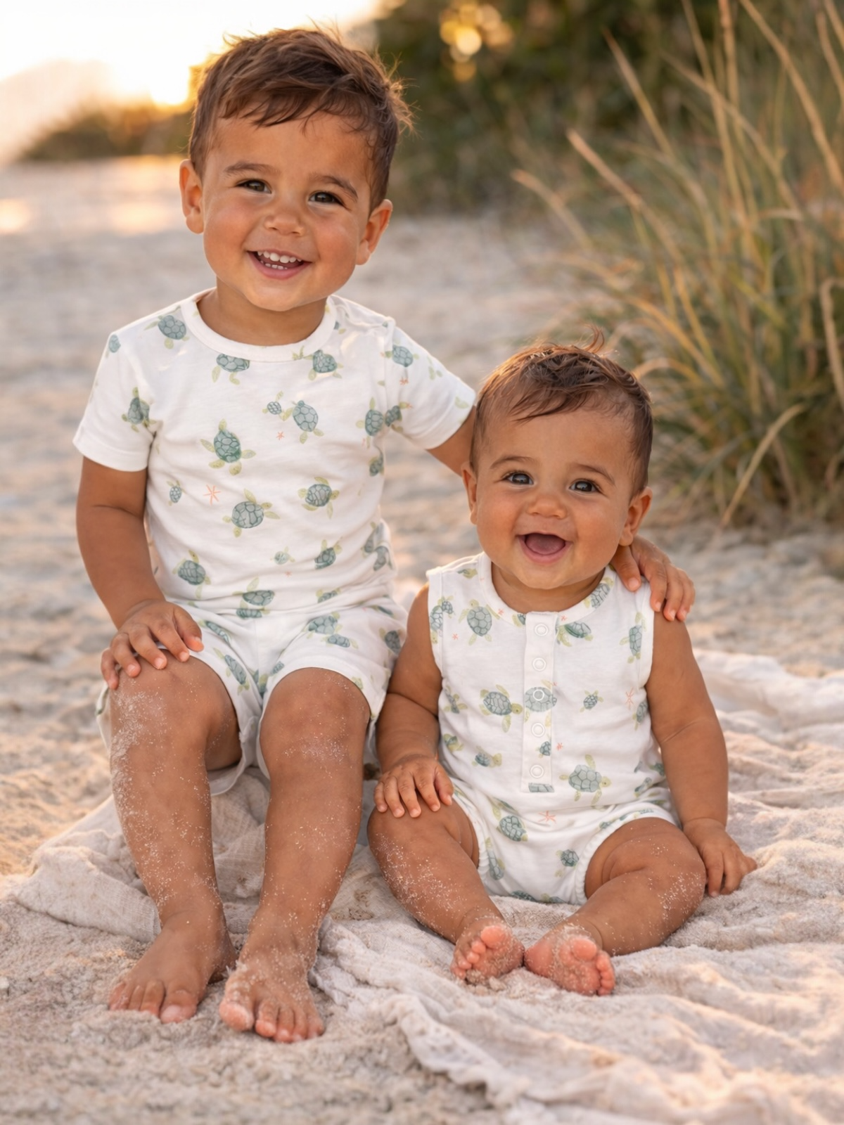 Two young boys in matching turtle-patterned outfits sit on a beach, smiling against a sunny backdrop.