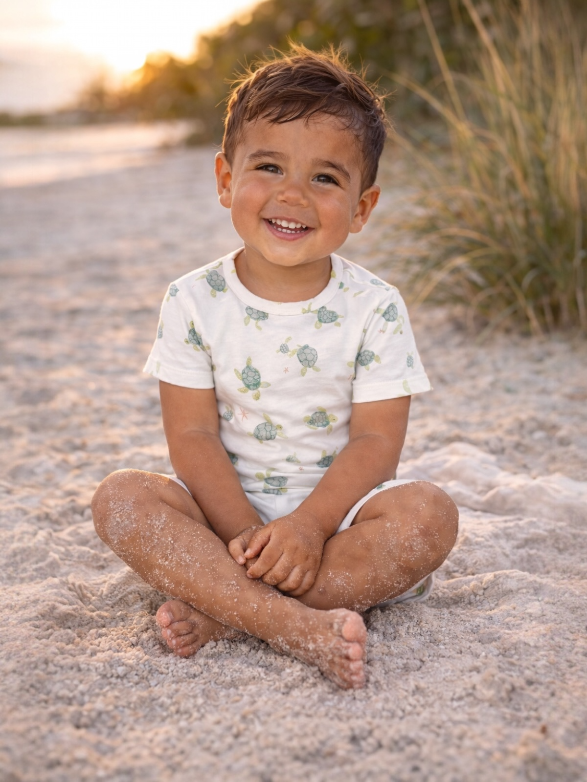 Smiling boy sitting cross-legged on sandy beach, wearing a turtle-patterned shirt, with sunlight in the background.