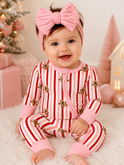 Smiling baby in festive striped pajamas with gingerbread design, pink headband, sitting on fluffy surface.