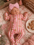 Baby in a striped gingerbread onesie and pink headband, resting on a pink blanket with holiday decor.