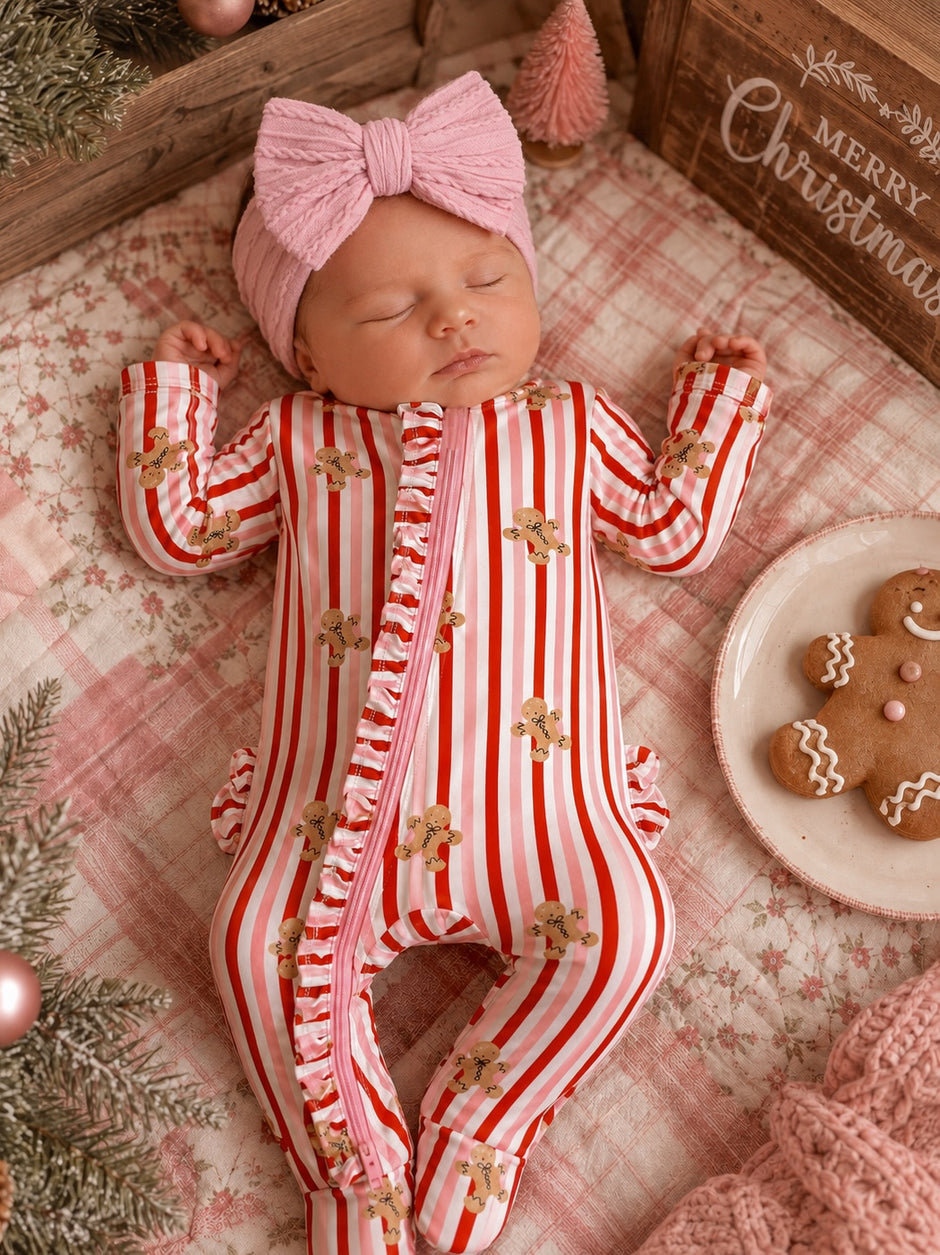 Baby in a striped gingerbread onesie and pink headband, resting on a pink blanket with holiday decor.
