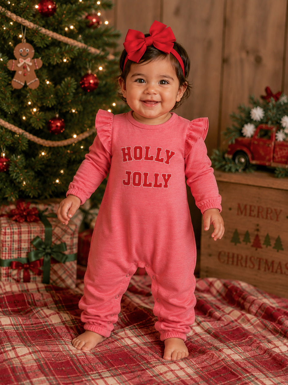 Smiling toddler in a pink jumpsuit with "HOLLY JOLLY" text, posed in front of a Christmas tree and decorated gifts.