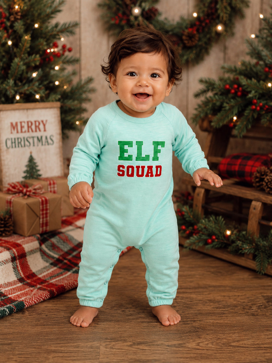 Smiling baby in a light blue onesie with "ELF SQUAD" text, in a festive holiday setting with decorations.