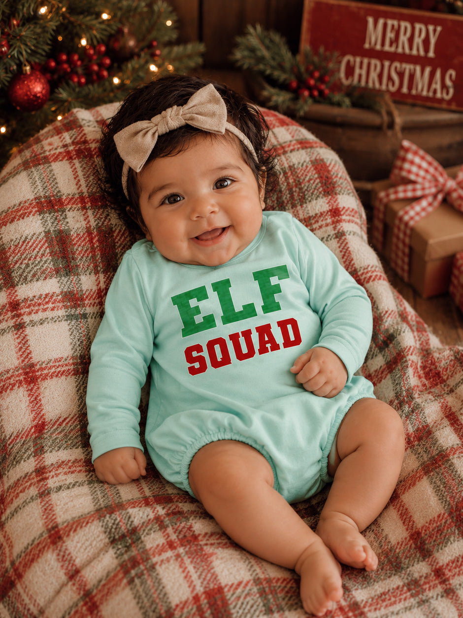 Smiling baby in mint outfit with "ELF SQUAD" text, sitting on plaid blanket near Christmas decor.
