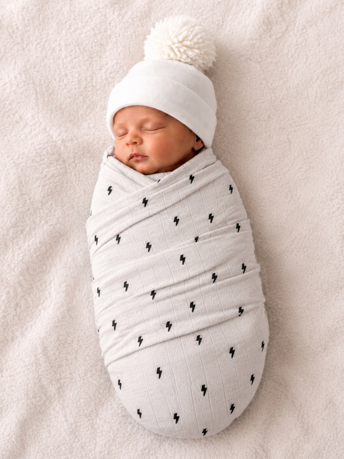 Newborn baby wrapped snugly in a light blanket with lightning patterns, wearing a white hat with a pom-pom.