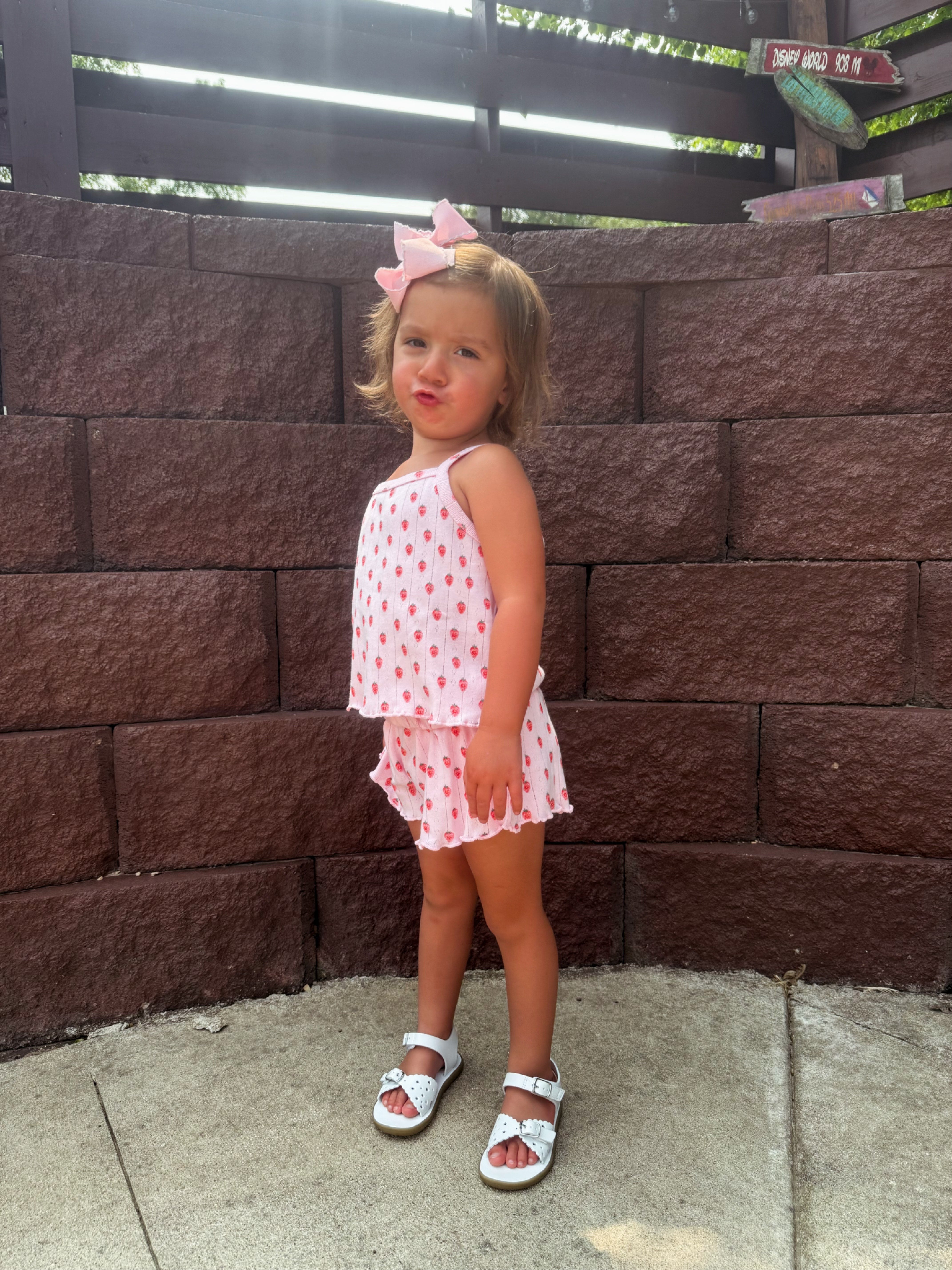 Young girl in a pink polka dot outfit and a big bow, standing on a sidewalk against a textured brown wall.