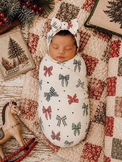 Infant swaddled in a bow-patterned blanket, resting on a quilt with holiday decor and a wooden rocking horse.
