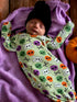 Newborn in a green skull print outfit, resting on a purple blanket next to a small pumpkin, wearing a black beanie.