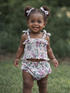 Smiling toddler in floral outfit stands in grass, wearing matching hair bows and displaying joyful expression.
