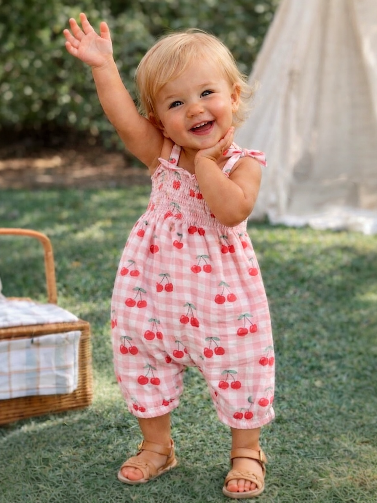Smiling toddler in a cherry-patterned romper waves happily outdoors. Green background with a picnic setup nearby.