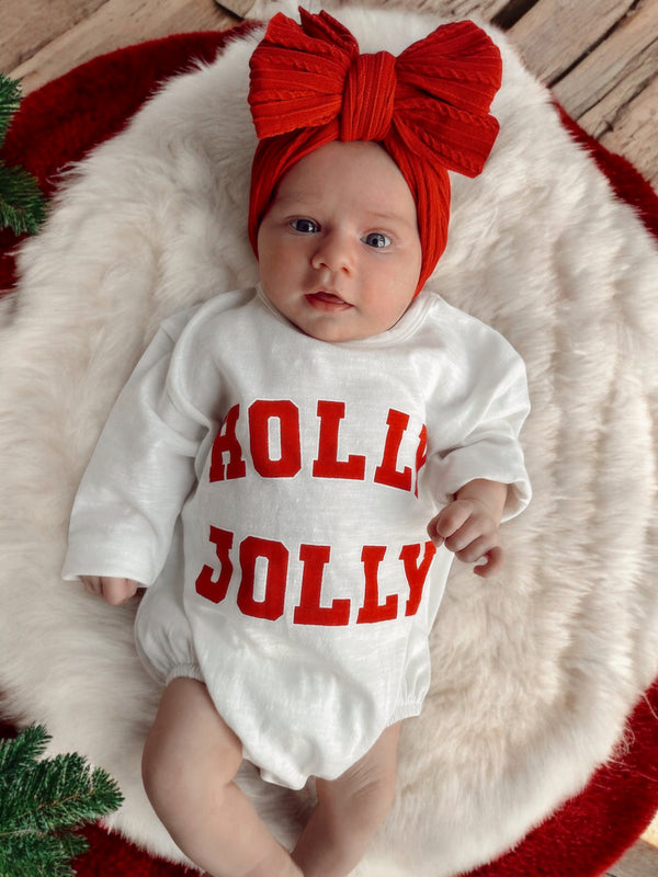 Baby wearing a white "Holly Jolly" outfit with a red bow headband, lying on a fluffy blanket.