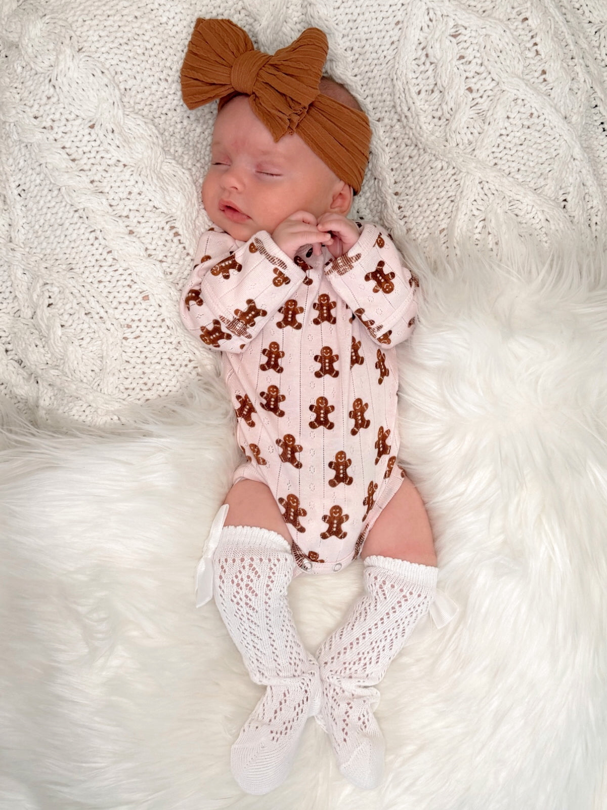 Baby girl in gingerbread onesie and bow, resting on white fuzzy blanket. Soft, cozy, and festive vibe.
