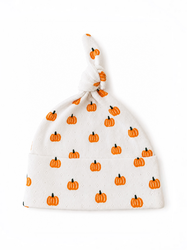 White baby hat with a knot, decorated with small orange pumpkin patterns.