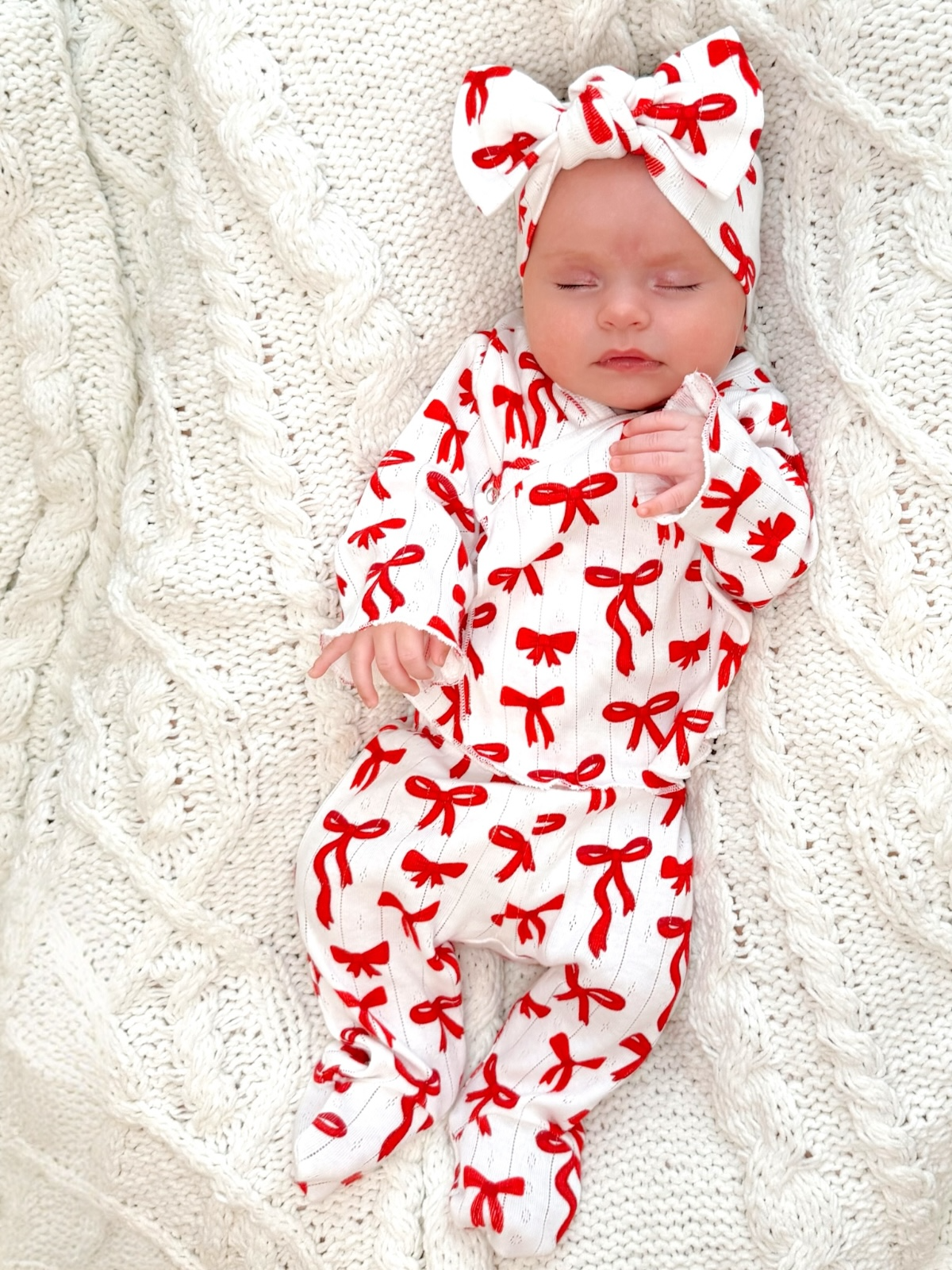 Baby girl in red and white bow-patterned pajama set, resting on a textured white blanket.