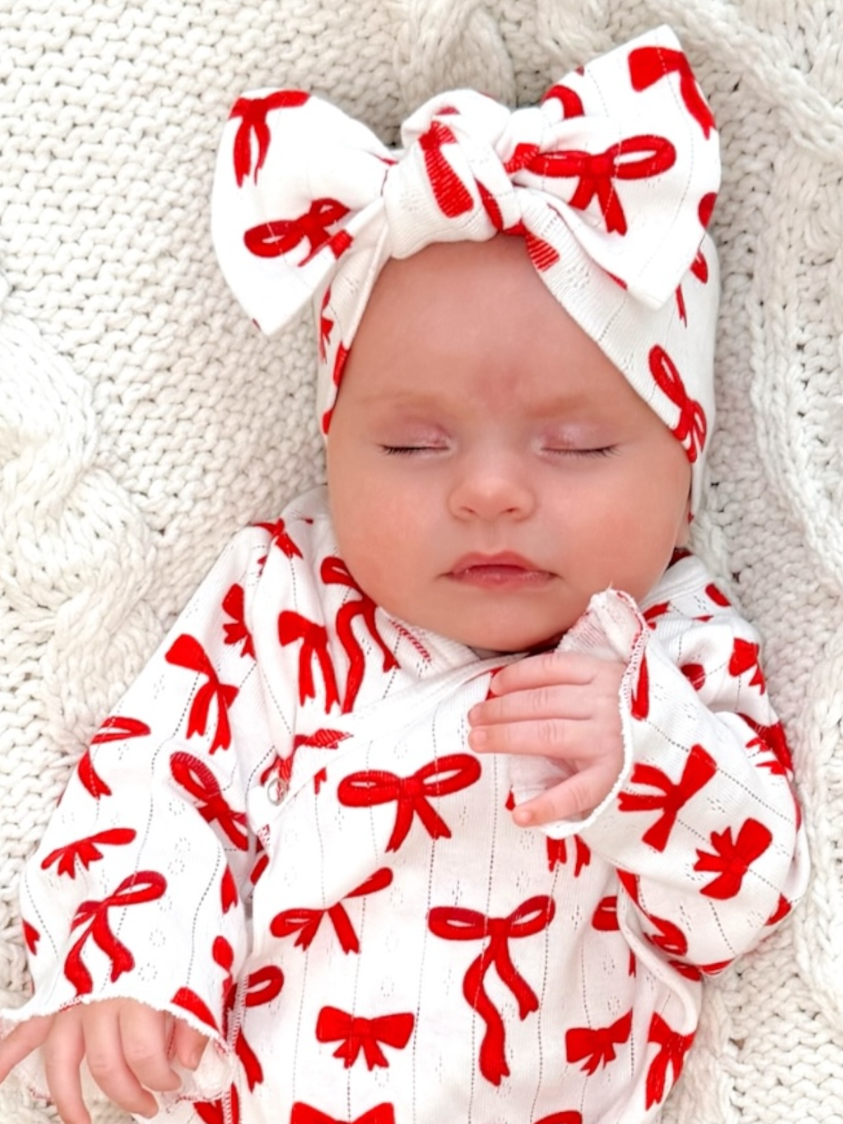 Baby girl peacefully sleeping in a cozy outfit with red bows, wrapped in a knit blanket.