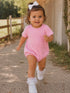 Smiling toddler in pink outfit running on a gravel path with greenery and wooden fence in the background.